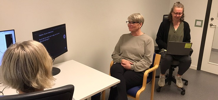 Vårdpersonal och patient med hörselnedsättning. Tolken sitter i rummet och tolkar till skriven svenska. Patienten kan läsa på skärmen vad vårdpersonal säger.